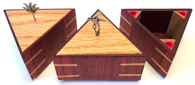 A photo of three wooden triangular boxes made from padauk wood, with oak highlights. Two of them have oak lids with silver sticking out of the top.