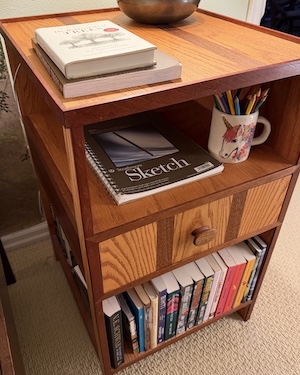 A photo of a nightstand bookshelf made from oak and jatoba. There are three sections. The top section is open, the middle section has a drawer, and the bottom is also open.