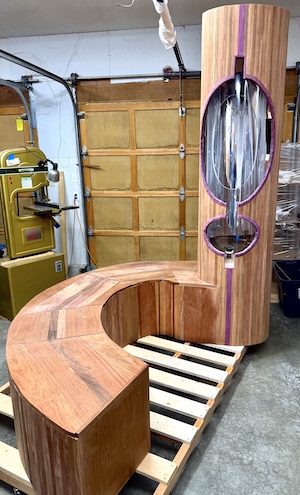 A photograph of the wooden frame for Rain Chamber #1. It is a vertial, cylndrical tower with an attached, curving bench. The tower has a large and small oblog-shaped cutout. The tower and bench is jatoba wood and the cutout is detailed with purplewood.